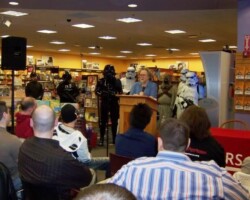 Aaron Allston stands at a podium in front of an audience with various Star Wars costumers behind him.