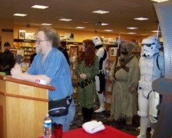 Aaron Allston leaning on a podium with Star Wars costumers behind him, shot from the right